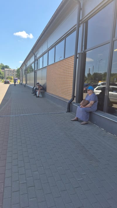 The image depicts a street scene in Preiļi, Latvia, likely on a sunny day given the bright blue sky and clear shadows. The main subject is a woman wearing a blue and white striped dress and a blue cap. She is sitting on the ground near the entrance of a modern building with large glass windows. She is leaning forward, with a tan purse slung over her shoulder.
In the background, under the overhang of the building, a group of three people are seated on the ground. They appear to be resting or waiting. The building has a reflective glass facade and a textured wall that appears to be made of small, square, beige tiles. The ground is paved with grey brick pavers, with a section of reddish-brown pavers visible in the foreground.
Further in the background, a few cars are visible on a road, and residential buildings can be seen amidst green trees. The overall atmosphere seems relaxed and casual, suggesting a typical day in a town. The time of day appears to be daytime, likely late morning or afternoon, due to the strong sunlight and distinct shadows. No visible text is present in the image.