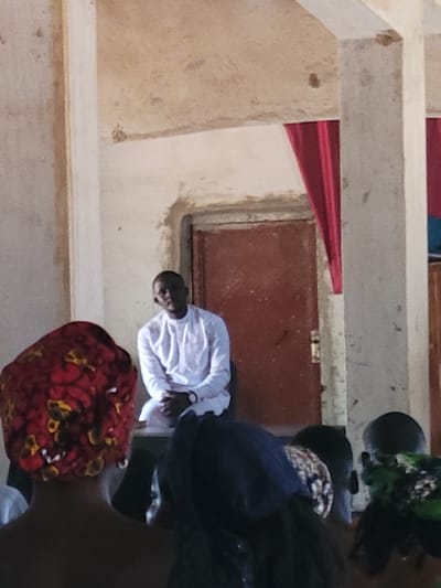 An indoor scene in Rinze, Nigeria, depicts a gathering. A dark-skinned adult male, wearing a long-sleeved white garment and a wristwatch on his left wrist, is seated in the mid-ground, facing generally forward. He is positioned in front of a reddish-brown door with visible hardware and a red curtain or drape on the right. The walls and ceiling are light-colored, exhibiting signs of wear and discoloration. Two light-colored concrete pillars are visible on the left and right sides of the frame. In the foreground, several individuals are visible from the back of their heads and shoulders, oriented towards the seated man. One person on the left wears a red, yellow, and black patterned fabric headwrap, and another in the center wears a dark head covering.