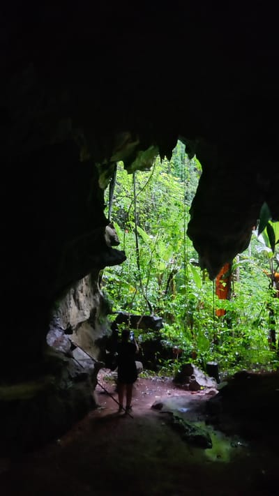 A dimly lit cave entrance frames a view of dense green foliage, likely located in Ban Pak Ra, Thailand. A person stands near the edge of the cave, seemingly using walking sticks. The interior of the cave is dark, revealing rough rock formations. The light streaming through the entrance illuminates the vegetation and suggests a tropical environment.