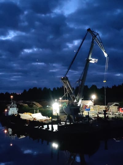 The image depicts a port scene in Annensky Most, Russia, at night. A large crane dominates the center of the frame, illuminated by several lights. The crane is positioned on a platform next to the water, with stacks of logs and lumber visible around it. A small tugboat is docked on the left side of the frame, and a truck is parked on the right. The sky is dark blue and cloudy, with a few visible stars. The water reflects the lights from the crane and surrounding structures, creating a mirrored effect.