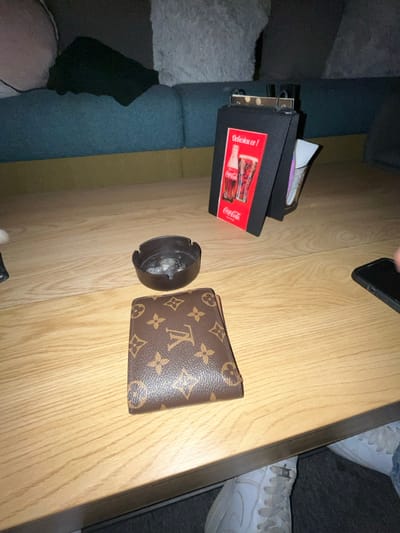 The image displays an indoor scene, likely a cafe or lounge in Пампорово, Bulgaria. The primary focus is a light-colored wooden table. On the table, from left to right, are a black, circular ashtray containing white ash residue, a closed brown wallet embossed with the Louis Vuitton monogram pattern, and a black table display stand. The stand features a red Coca-Cola advertisement showing a bottle and a glass, with text including "Delicious!" and "Coca-Cola". Behind the stand, a clear glass container holds several white napkins. A black smartphone is partially visible on the far right edge of the table, possibly being held or interacted with by a person whose hand is just out of frame. In the background, a section of a blue upholstered sofa is visible, with a light grey cushion or throw. Under the table on the right side, a person's lower leg, clad in light blue jeans, and a white athletic sneaker are visible, resting on a dark grey floor surface.
