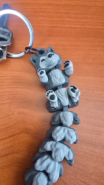 A close-up overhead shot shows a string of gray wolf figurines hanging from a silver keychain. The wolf figurines are connected in a chain, with each wolf in a slightly different pose. They are lying on a wooden surface with a visible grain. The keychain itself has a large ring and appears to be attached to a small object, possibly a key. The lighting suggests an indoor setting. This image is not located in Krasnopol’ye, Russia.