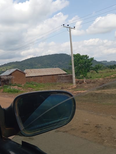 A rearview mirror of a vehicle shows a rural landscape in Jibam, Nigeria. In the distance, two mud-brick houses with corrugated metal roofs are visible, situated in a clearing with green grass and some scattered debris. A tall, thin utility pole with wires stretches across the frame. Behind the houses, a gently sloping, tree-covered hill forms the horizon under a sky with scattered white clouds. A lone tree with dense green foliage stands to the right of the utility pole, and a small herd of animals, possibly goats, can be seen grazing in the middle ground. The foreground is dominated by the rearview mirror and the road surface, which appears to be dirt.