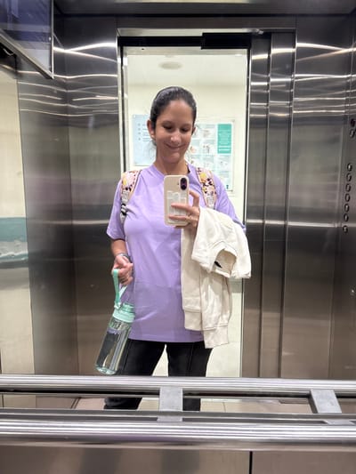 A woman is taking a selfie in an elevator. She is wearing a purple shirt, holding a water bottle in one hand and a cell phone in the other. She is also holding what appears to be a folded light-colored garment. The elevator has stainless steel walls and a metal handrail. The reflection shows a white wall with a sign on it. The location is likely not La Victoria, Peru, as there is no way to know this from the image.