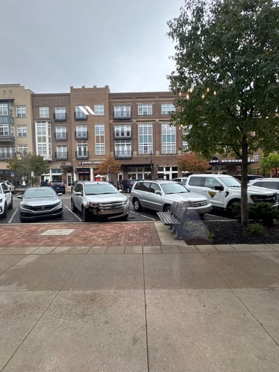 This image depicts a daytime urban scene under an overcast sky at Eddy Street Commons in South Bend, Indiana, United States.
In the foreground, a light-colored concrete sidewalk transitions to a section of reddish-brown brick pavers. To the right, a dark slatted bench is partially obscured by a tree with green foliage, within which string lights are visible. The base of the tree is surrounded by a mulched planting bed.
The midground features a parallel parking area. Visible vehicles include a silver Honda Civic sedan, a silver Ford Edge SUV exhibiting significant damage to its front bumper on the driver's side, a silver Toyota Highlander SUV, and a white Ford F-150 pickup truck with a yellow decal on its rear passenger door. Numerous other vehicles are partially visible further back in the lot.
The background consists of a row of four-story brick buildings, characterized by light-colored window frames and multiple balconies with dark railings. Ground-level retail spaces are present, with "Jimmy John's" and "JRU BURGER" signs clearly visible. A prominent white geometric logo, resembling a stylized 'W' or 'M', is affixed to the facade of the central building. No discernible individuals or specific actions are visible within the frame.