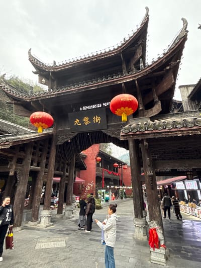The image displays an outdoor scene at Jiuli Street in Pengshui Miao and Tujia Autonomous County, China. The foreground is dominated by a large, dark-stained traditional Chinese archway, characterized by multiple tiers of curved tiled roofs and numerous substantial wooden support pillars resting on stone bases. A black rectangular sign with golden Chinese characters "九黎街" (Jiulijie) is centrally suspended from the archway. Two round red lanterns with golden patterns and yellow tassels hang from the archway, one on each side of the sign. Small string lights are visible hanging beneath the archway structure.
Beyond the archway, a paved pedestrian street extends into an area lined with traditional multi-story buildings, some featuring prominent red-painted walls and dark roofs. These buildings are decorated with additional red lanterns and red fabric streamers. Green, tree-covered hills or mountains are visible in the background.
On the left side of the foreground, a woman in a black jacket with white lettering and dark pants holds a yellow and black striped shopping bag, her mouth slightly open. To the right, another woman, dressed in a light-colored jacket and blue jeans, with her hair in a bun, faces right and extends her right arm, pointing forward. In the mid-ground, under the archway's left side, a group of three individuals appears to be conversing. Further down the street on the right, additional people are visible, including one man in a striped shirt. The sky is overcast. A small stone lion statue with red ribbons is present at the base of a right pillar.
