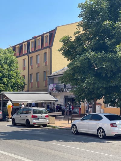 The image depicts a street scene in Stupava, Slovakia, on a sunny day. In the foreground, a white Skoda Octavia sedan is parked on the right side of the road. To the left of the sedan, a silver Volkswagen Touran is parked. Both vehicles are facing away from the viewer.
Behind the parked cars is a row of establishments. A large, oversized ice cream cone sculpture stands prominently on the left, in front of an awning that reads "SPECIAL GELATO". People are seen queuing and standing at the gelato shop. Adjacent to the gelato shop is another building with signage that includes the word "GELATO".
Further back, a multi-story building with a yellow facade and a mansard roof is visible. The windows of this building have red and yellow accents. Lush green trees with dense foliage line the street and partially obscure the buildings. The overall environment suggests a commercial or residential area with pedestrian activity. The sky is clear and blue.