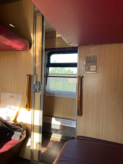 The image shows the interior of a compartment on a train. There are two bunk beds, one on either side of the compartment, upholstered in burgundy. A small window with a white curtain is visible. Above the window is a control panel with various buttons and a power outlet. There's also a folding table, part of which is visible on both sides of the window. A white plastic bag, and what appears to be clothing, are on the table on the bottom bunk. The location is Заслаўе, Belarus.