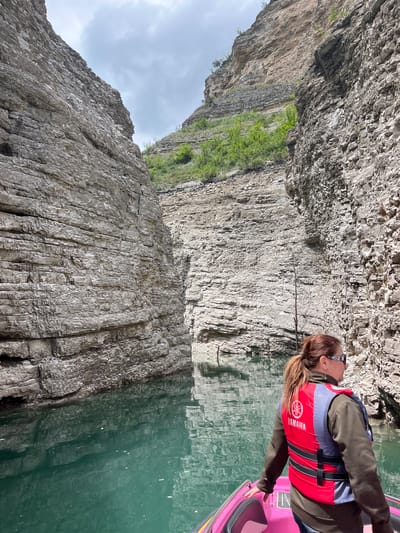 A woman wearing a red and blue life jacket sits in a pink kayak on a body of water. Towering rock formations line the water's edge. The sky is overcast. The brand "Yamaha" is visible on the life jacket. A city of Druzhba, Russia, location cannot be confirmed.