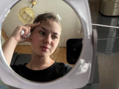 A young Caucasian female is reflected in a mirror, indoors. She has fair skin with freckles and some acne visible on her forehead and cheeks. Her dark blonde hair is pulled back, with a clear plastic hair clip visible at the top of her head. She is wearing a black ribbed t-shirt and a thin silver chain necklace. Her right hand is raised to her forehead, and she is holding a small piece of white tissue in her fingers. Her fingernails are painted black and white. The background reflected in the mirror shows a wall with cork tiling, a light fixture on the ceiling, and other objects that are out of focus. The mirror itself appears to be a portable vanity mirror with a light purple frame, which is partially visible in the foreground of the image. The overall impression is that of a personal moment of self-examination or skincare routine.