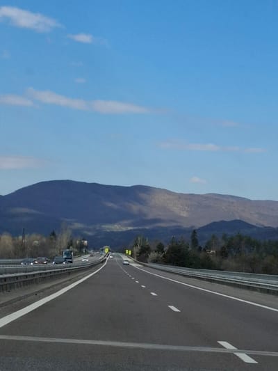 A highway with multiple lanes is visible, lined with a guardrail. Traffic is present, with several vehicles moving along the road. To the left of the highway is a large mountain range. The sky is blue with some streaks of light clouds. The picture may have been taken near Церово, Bulgaria.