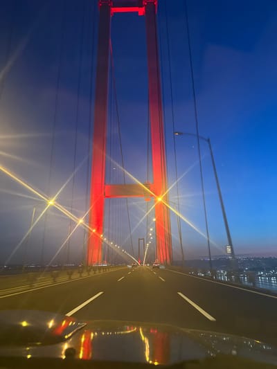The image depicts a bridge at night in Dilovası, Turkey. The bridge's support structure is painted red and illuminated. There are overhead lights along the bridge, and the roadway has lane markings. The image was taken from a vehicle on the bridge, with reflections of the bridge lights visible on the vehicle. The sky is dark blue, and city lights are visible in the background.