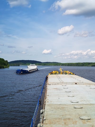 The image shows a wide river scene in what is likely the city of Gorka, Russia. Two large cargo ships are visible on the water. One ship, positioned closer to the viewer, has a flat, grey surface with circular hatches and yellow superstructures. The second ship, further in the distance, is blue and white with a cabin. A blue railing lines the edge of the first ship. The river is a dark, rippling blue. Both sides of the river are lined with green trees and hills. The sky is light blue with scattered white clouds.