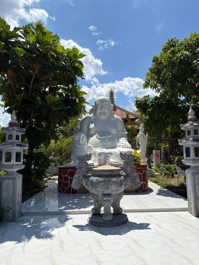 A large, white, seated Buddha statue is the central focus of the image, positioned outdoors in what appears to be a temple courtyard. The statue is adorned with a beaded necklace and rests on a circular base with red and white patterned tiling. In front of the Buddha statue is a large, ornate, grey stone incense burner, with two stylized dragon heads on either side. Flanking the scene are two white, multi-tiered pagoda-like structures.
Behind the Buddha statue, a smaller white statue of a standing female figure is visible, along with a stone pillar bearing red Chinese characters. The background features lush green trees with large leaves, suggesting a tropical or subtropical climate. A building with a terracotta roof and a tiered spire is partially visible behind the trees. The ground in the foreground is tiled with large, white marble-like slabs, reflecting sunlight and casting shadows from the surrounding objects and foliage. The sky above is a clear blue with scattered white clouds.
The scene is likely located in a temple or religious complex in Tuy An Nam, Vietnam, given the architectural style and religious iconography. The overall impression is one of serene tranquility, typical of such spiritual sites.