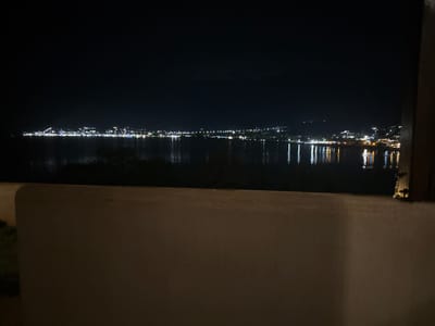 This image captures a night scene of the Skala Potamias coastline in Greece. The foreground features a light-colored, textured wall with a slight curvature, suggesting it's part of a balcony or viewing area. Beyond the wall, a dark body of water, likely the sea, stretches across the frame. The distant shoreline is illuminated by numerous lights, indicating a town or village. The lights are concentrated along the coast, with some appearing on higher ground, suggesting a hilly terrain. The reflection of these lights shimmers on the calm surface of the water. The sky above is dark, indicating nighttime. The overall impression is one of a peaceful, illuminated coastal town at night.