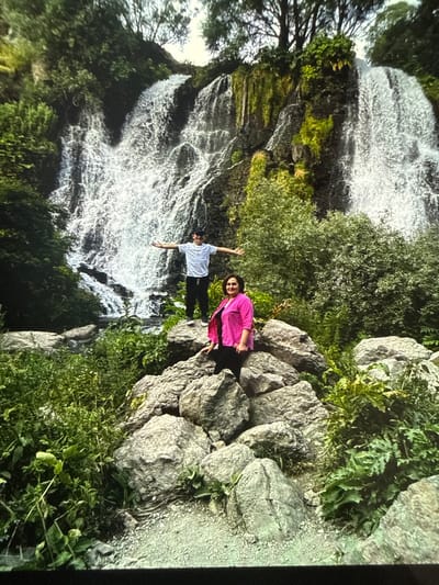 The image captures a scenic waterfall in Artik, Armenia. In the foreground, a woman in a bright pink shirt and black pants is seated on large rocks, smiling. To her left, a young man in a white t-shirt and black pants stands on a rock with his arms outstretched, facing the waterfall. The waterfall itself is majestic, with water cascading down a rocky cliff face covered in greenery. Lush vegetation surrounds the waterfall and the rocky outcrop where the two individuals are positioned. The overall scene depicts a serene natural environment.
