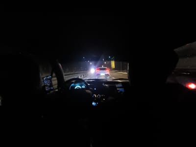 The interior of a car at night. The illuminated dashboard is visible, as is the steering wheel. Through the windshield, another car can be seen on the road ahead, along with streetlights and roadside structures. Two figures are silhouetted in the front seats. One person is using a smartphone. The overall lighting is dim, creating a sense of nighttime travel.