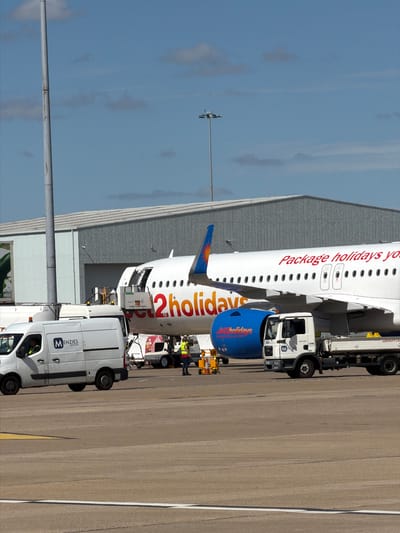 This is a photograph of an airplane parked at an airport, likely Luton Airport in the United Kingdom, as indicated by visible text. The plane, which appears to be a passenger jet, has "Package holidays yo" and "2holidays" written on its side in red lettering. A blue section on the front of the plane also has "2holidays" written in white.
Several service vehicles are positioned around the aircraft. One vehicle, a white van with "MENZIES AVIATION" on its side, is prominent in the foreground. Another, a white truck with a yellow striped trailer, is closer to the airplane's wing. There are also workers visible near the airplane and the vehicles, suggesting ground operations are underway.
The scene appears to be set during the daytime, under a clear blue sky with some wispy clouds. The sun is casting shadows, indicating it is likely a bright and sunny day. The airplane is parked on a tarmac, with a large hangar-like building in the background. The overall impression is one of an active airport environment, with aircraft being serviced between flights.