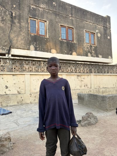A young boy stands centrally in an outdoor setting in Daurawa, Nigeria. He is approximately 8-12 years old, with dark skin and short, dark hair. He wears an oversized navy blue V-neck sweater with thin red stripes on the collar and cuffs, and a small circular gold emblem on the left chest. He also wears dark grey trousers. His left hand holds a black plastic bag, which hangs at his side. The boy gazes directly forward with a neutral expression.
Behind the boy, a weathered two-story building with a grey-beige, textured facade showing signs of wear and discoloration dominates the background. The upper story features three rectangular windows with brown wooden frames and translucent blue panes; two are visible on the main face and one on a recessed section to the left. Below the main structure, a light-colored wall or fence extends horizontally, decorated with a repeating pattern of geometric perforations. An electrical cable runs vertically down the left side of the building.
The ground surface is a mixture of light brown, granular soil/sand and irregular concrete paving slabs, particularly visible beneath the boy and to his left. To the immediate left of the boy, a folded blue fabric item rests on the ground. To his right, a large, dark, irregular rock-like object is present, with a low, rectangular concrete structure visible behind it. The upper right portion of the frame shows a clear, light blue sky.