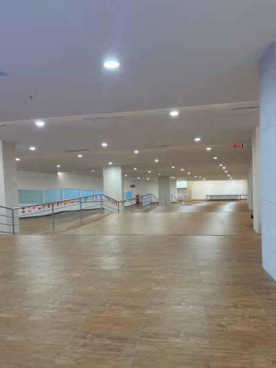 The image depicts an indoor, well-lit hallway or public space with a polished wooden tile floor. The ceiling features multiple round lights and rectangular fluorescent fixtures. A ramp with a metal railing is visible on the left, adorned with a string of small international flags. Further down the hallway, a set of stairs with a similar railing is also present. In the distance, a row of shopping carts is neatly arranged against a white wall. Several large windows with light blue panes line the upper portion of the walls. The environment appears to be a modern public building, possibly a transportation hub or a large commercial establishment, located in Baji Mangngai, Indonesia. There are no people visible in the scene.