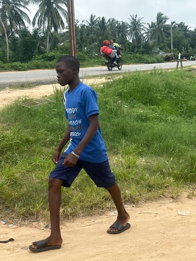 The image depicts a young Black male walking on a dirt path adjacent to a paved road lined with lush green vegetation and palm trees. He is wearing a blue t-shirt with white lettering that reads "NOBODY KNOWS TOMORROW" and dark blue shorts. On his left wrist, he wears a silver chain bracelet. He is also wearing black slide sandals. In the background, a motorcycle carrying multiple people and cargo travels along the paved road. The sky is overcast. The scene appears to be in Ganyingbo, Nigeria, a coastal area known for its palm trees and tropical vegetation.