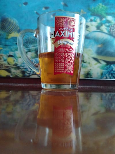 A glass mug filled with amber-colored liquid, likely tea or coffee, rests on a reflective wooden surface. A red and gold label with ornate patterns and text indicating "COFFEE MAXIMUS Original FREEZE-DRIED" is adhered to the mug. The mug has a clear handle. The background features a blurred image of underwater life, possibly a poster or aquarium scene, with fish and coral-like structures in shades of blue, green, and yellow. The lighting suggests an indoor setting. The image was captured in Geologorazvedka, Russia.