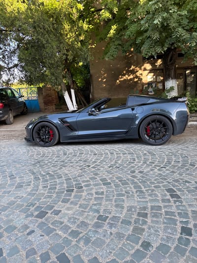 The image shows a gray convertible sports car parked on a cobblestone street in Hayredin, Bulgaria. The car is parked next to a building with peeling paint and trees growing nearby. The car's door is open. A glimpse of another car is visible behind the sports car. The cobblestone street is a gray color. The image is taken in daylight.