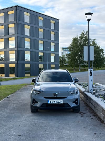 In Östersund, Sweden, an electric Volvo car is parked on a concrete surface next to a modern grey building with golden-trimmed windows. A charging station is attached to the car, and a street lamp stands nearby. Trees are visible in the background, along with a building that appears to be an air traffic control tower. The sky is partly cloudy.
