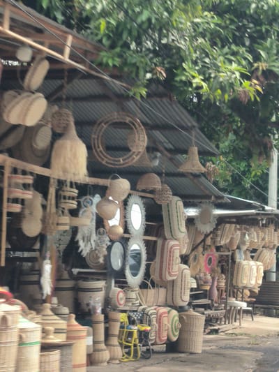 The image depicts an outdoor marketplace stall in Aneuk Paya, Indonesia, showcasing a variety of handcrafted woven goods. The stall is constructed with a corrugated metal roof supported by wooden beams. Hanging from the roof and displayed on shelves are numerous baskets, bowls, lampshades, and decorative circular items, all made from natural woven materials like rattan or bamboo. Many of these items are light-colored, with some featuring subtle red or green accents. In the foreground, a collection of large, cylindrical baskets are arranged. A yellow metal chair with a red seat and green accent is visible to the right, and further in the background, a wooden bench can be seen. Lush green foliage from trees overhangs the stall from above, providing shade. The ground in front of the stall appears to be a paved surface. No people are visible in the frame.
