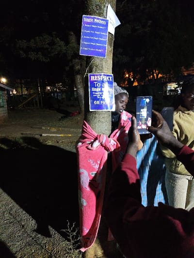 A gathering takes place at night around a tree adorned with signs. The signs are advocating for single mothers, stating "Respect Single Mothers", and detailing a 68-hour "Hugging to All Single Mothers" event. A woman stands next to the tree wrapped in a pink blanket. A person holds up a phone, apparently to record the scene, while another individual stands nearby. The overall scene conveys a message of support and advocacy for single mothers.