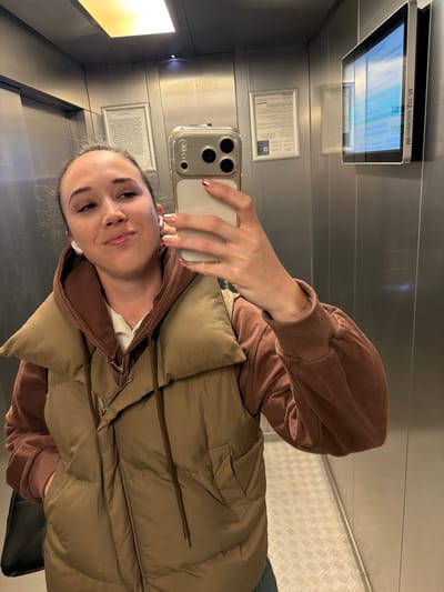 The image shows a woman taking a selfie in an elevator. She is wearing a brown puffer vest over a brown long-sleeved top and has white earbuds in her ears. Her phone is silver with multiple lenses. There is a digital display screen above and behind her, along with informational signage on the wall. The elevator has metal walls and a textured metal floor. The city of Leninavan, Russia is not evident in the image.