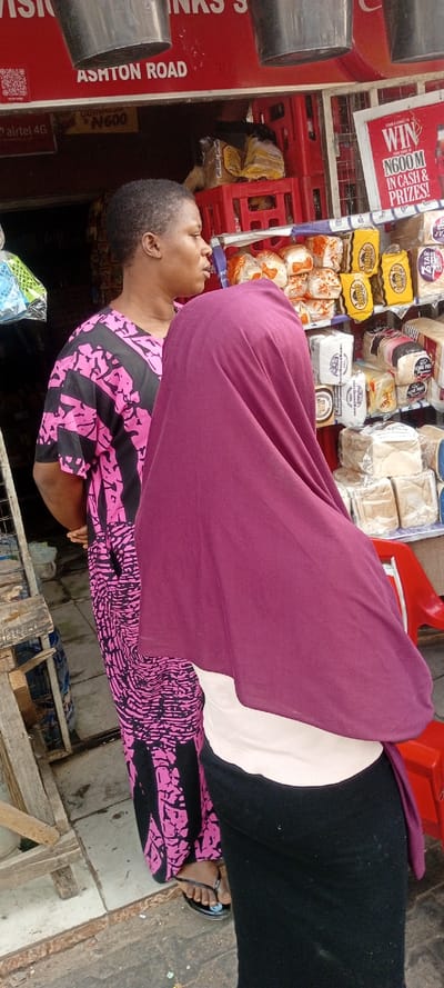 Two women stand near a roadside vendor. One woman wears a pink and black patterned dress and black sandals. The other woman wears a black skirt, white top, and a large maroon headscarf that obscures her face. The vendor's stall displays various loaves of bread. A red sign advertises the chance to win money and prizes. The sign above the vendor's stall includes the words "Ashton Road".