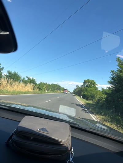 The image, taken from inside a vehicle, shows a sunny day with a clear blue sky. The car is driving on a paved road lined with trees and dry grasses. In the distance, there are a few other vehicles on the road, including a red truck. The car's dashboard is visible in the foreground, with a dark grey bag on it. The scene appears to be in a rural or suburban area, possibly along a highway. The location is specified as being in the city of Vinitsa, Bulgaria.
