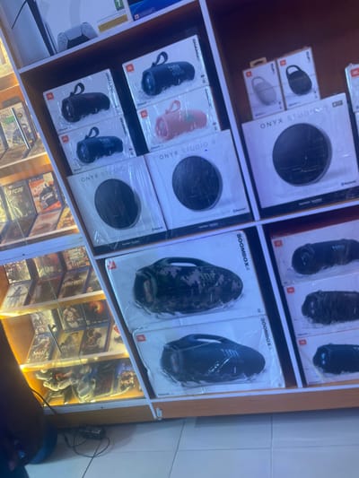 The image shows a display shelf in a retail store, possibly in Ugbor, Nigeria, based on the OCR. The shelf contains several product lines, including JBL Charge speakers, Onyx Studio speakers, and JBL Boombox speakers. Additionally, there's a display of PlayStation 4 (PS4) games. There are a pair of white PlayStation 5 controllers. The shelf is brightly lit, and the items are neatly organized. The image also features a blurry view of the lower legs of a person standing in front of the shelf.