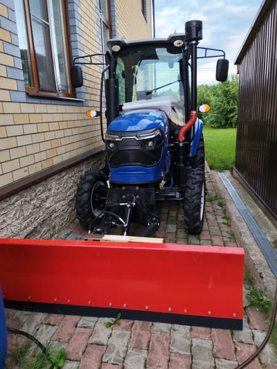 A blue tractor with a red snowplow attachment is parked on a paved driveway in front of a brick building and a dark metal fence. The tractor has large, rugged tires, a cab with windows, a prominent exhaust pipe, and headlights. The snowplow is wide and bright red, with a black rubber blade along the bottom. The building appears to be a house or garage with a window visible. The ground is covered in red and grey paving stones. Green grass and bushes are visible in the background. The weather appears to be overcast. The location is identified as Kstovo, Russia.