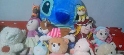 This media file displays a collection of plush toys arranged on what appears to be a sofa or bed. The setting is indoor, with a plain blue wall in the background and a red patterned pillow or blanket visible in the foreground. The toys are diverse, including a large blue Stitch character from Disney's Lilo & Stitch, several teddy bears in various sizes and colors, a white bear with a red apple design on its chest, a dog plush wearing an orange and blue uniform, another dog plush wearing a yellow hat and uniform, a doll with blonde yarn hair and a red dress, and a doll with pink yarn hair and a blue outfit. There is also a light blue plush toy with a unicorn-like appearance. The lighting suggests daytime, but no specific time of day can be determined. There is no indication of weather or a specific location beyond "indoor." The toys are positioned in a static arrangement, implying no particular activity or event is taking place. The overall mood is playful and childlike, as expected from a collection of stuffed animals. No visible text is present in the image.