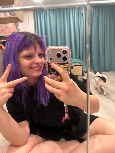 A woman with vibrant purple hair smiles and makes a peace sign while taking a mirror selfie with her phone. The phone case is clear with a pink border and a colorful design. She is wearing a black shirt. In the background, a blue curtain hangs behind a small wooden bedside table. A black and white dog lies on the floor near the curtain.