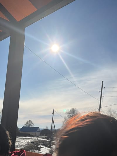The image shows a bright, sunny sky with the sun prominently visible. A utility pole with power lines stretches across the upper portion of the image. The roof of a structure is visible in the bottom left corner. In the background, a house with a dark roof stands among snow-covered ground. A person's head is partially visible in the upper right and lower right corners. The location is consistent with a rural setting in Gory, Russia, given the presence of utility poles and residential buildings.