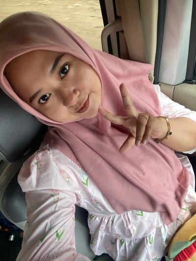 A woman wearing a pink headscarf is seated in a car. She is positioned at an angle, looking directly at the camera with a smile. Her right hand is raised, displaying a peace sign near her face. She wears a patterned, light-colored outfit with ruffles. A gold bracelet adorns her wrist. The interior of the car is visible, including a gray seat and a door handle. A hamburger plush is seen in the background.