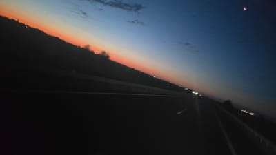 The image depicts a highway scene at dusk, with a clear view of the horizon transitioning from a deep blue sky to a gradient of orange and pink. The road appears to be a multi-lane highway, with white lane markings visible. There are faint lights of distant vehicles and structures along the side of the road. A small, crescent-shaped object, possibly the moon, is visible in the upper right corner of the sky, tinged with pink. The overall impression is one of a peaceful journey at the end of the day, with the urban landscape of Kövegy, Hungary, dimly visible in the distance.