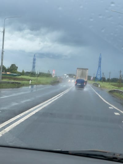 The image, taken from inside a vehicle, shows a wet road during a rainy day in Кокино, Russia. A truck and a blue car are visible ahead on the road. Power lines and utility poles run along the side of the road. A gas station sign is also seen. Raindrops are visible on the windshield. The weather is overcast. The location is likely a highway or major road due to the presence of dividing lines.