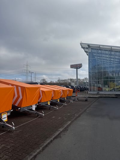 This image depicts the exterior of an OBI store in Gießen, Germany, under an overcast sky. The foreground features a linear arrangement of approximately ten small utility trailers, each covered with a bright orange tarpaulin displaying a white chevron pattern pointing right. The trailers are secured by chains connecting their hitch couplers. A yellow-red price tag on the leftmost trailer indicates "AKTION" and a price of "549,-". The trailers are positioned on a paved surface composed of dark grey interlocking bricks, separated by a concrete curb from a darker asphalt parking area. In the midground, a two-wheeled hand truck with an orange handle is visible on the asphalt near the store entrance. Beyond the trailers and hand truck, a parking lot contains multiple dark-colored vehicles. The background is dominated by a large, modern OBI store building featuring an extensive glass facade, reflecting the cloudy sky. A tall pole sign displaying the "OBI" logo in red and white is located between the parking lot and the building. Further in the background, a line of deciduous trees without foliage suggests a cold season, and multiple high-voltage electricity pylons with power lines stretch across the horizon. No individuals are visible in the scene.