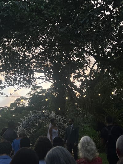The scene depicts an outdoor gathering, identified as taking place in Camaragibe, Brazil, during the late afternoon or early evening. The ambient light indicates dusk, supplemented by approximately fifteen spherical string lights suspended across the setting.
In the midground, two central figures are positioned. A female, presumed to be a bride, wears a white strapless gown and a white veil, and has dark hair. She stands to the left of a male, presumed to be the groom, who is dressed in a dark suit, light shirt, and tie, and has facial hair and eyeglasses. They stand before a white floral arch, which is set against a backdrop of dense green foliage.
The foreground contains the blurred heads and shoulders of numerous individuals, facing towards the central figures, indicating their role as attendees. Hair colors among these individuals vary, including dark, grey, and blonde. On the far right, a person in a dark shirt and backpack is visible, partially facing away.
The environment is characterized by a dense canopy of large, mature trees with dark foliage, silhouetted against a sky showing gradients of light and faint coloration, consistent with sunset or twilight. Further green foliage is visible throughout the background.