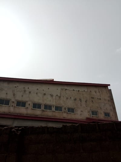 The media file shows the exterior of a building with a weathered, concrete facade. The building has a flat roof with a red overhang. Several rectangular windows are visible, evenly spaced along the building's wall. The facade is marked with numerous circular indentations, suggesting damage or an unusual construction feature. A dark, textured wall or fence runs across the foreground, obscuring the lower portion of the building. The sky is overcast and bright, indicating daytime. The scene appears to be an outdoor setting in Mbar, Nigeria. No people or specific activities are depicted. The overall impression is one of a functional, industrial or commercial structure, possibly showing signs of age or past events. There is no visible text in the image.