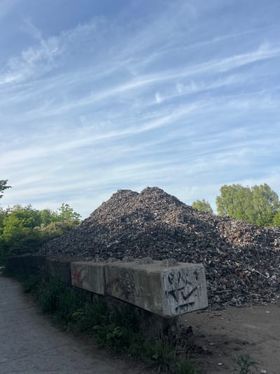 The image shows a large heap of scrap metal or waste material adjacent to a concrete barrier. The barrier appears to be marked with graffiti. The sky is visible with streaks of clouds. Vegetation, possibly trees, is visible behind and to the side of the scrap pile. The scene is located in Marquette-lez-Lille, France. No people are visible. A grey ground or pavement is in the foreground of the photo.