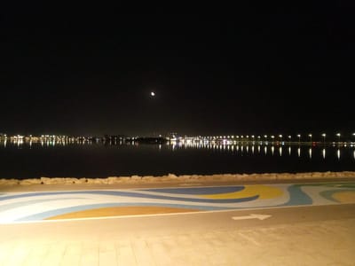 The image depicts a nighttime scene of a waterfront promenade in Al Khobar, Saudi Arabia. The foreground features a paved walkway with a colorful, wavy pattern in shades of blue, white, and yellow, possibly indicating a cycling or pedestrian path. A white arrow is painted on the path, pointing to the right. Beyond the walkway, a line of rocks separates it from a calm body of water, likely the Arabian Gulf. The water reflects the lights of a distant city skyline, characterized by numerous illuminated poles or lampposts that stretch across the horizon. A sliver of a crescent moon is visible in the dark night sky above the city lights. The overall atmosphere is serene and illuminated by artificial lights.
