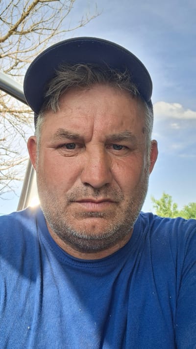 This media file is a portrait of a middle-aged man in Hîncești, Moldova. He is wearing a blue t-shirt and a dark blue baseball cap. The image is taken from a low angle, looking up at the man. His face is the primary focus, with his eyes looking directly at the camera. He has graying hair and a beard. The background consists of a bright blue sky with some clouds, bare tree branches, and a hint of green foliage, suggesting an outdoor setting possibly in late autumn or early spring. A bright sun flare is visible in the lower right portion of the image. The man's expression appears neutral or slightly serious. There is no visible text in the image.