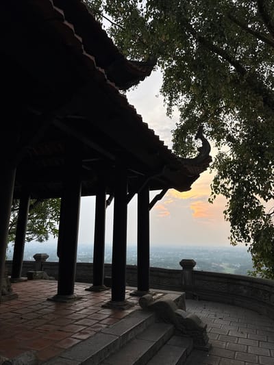 This is a medium shot, eye-level photograph of a pagoda-like structure in Phong Châu, Vietnam. The pagoda's dark wooden roof is in the foreground, casting a shadow over a tiled patio. The roof has ornate, upward-curving eaves with decorative elements. Several dark, thick pillars support the roof. The patio is made of terracotta tiles and transitions to stone steps. A stone balustrade with intricate carvings runs along the edge of the patio, overlooking a hazy city or town in the distance. Lush green trees with dappled sunlight filter in from the right side of the frame. The sky is a soft gradient of orange, yellow, and white, suggesting either sunrise or sunset. No people are visible in the image.