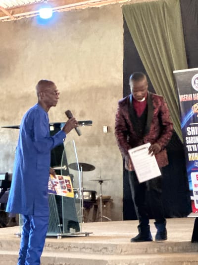 In Kufana, Nigeria, a man wearing a blue traditional outfit and glasses stands to the left, holding a microphone in his right hand and a brochure with images in his left. He appears to be speaking. To his right, a man in a dark suit with a red patterned jacket and a white clerical collar stands, holding a document. He is looking down at the document. In the background, a drum set and other musical equipment are visible. A large banner with text is positioned to the far right. The setting appears to be an indoor event, possibly a church or community hall, with a light fixture overhead.