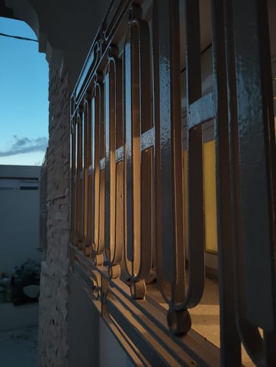 The image shows a close-up view of a decorative metal railing, likely a balcony or fence, in front of a stone-tiled wall. The railing has a repeated arched design. Sunlight illuminates the railing from the right, casting shadows and highlighting its texture. Behind the railing, a yellow light is visible, suggesting an interior space or a lit object. To the left, a clear blue sky with a few wispy clouds can be seen above the stone wall, which appears to be part of a building. The overall scene is likely outdoors, possibly at dusk or dawn, given the interplay of light and shadow. The location is identified as Ariana, Tunisia.